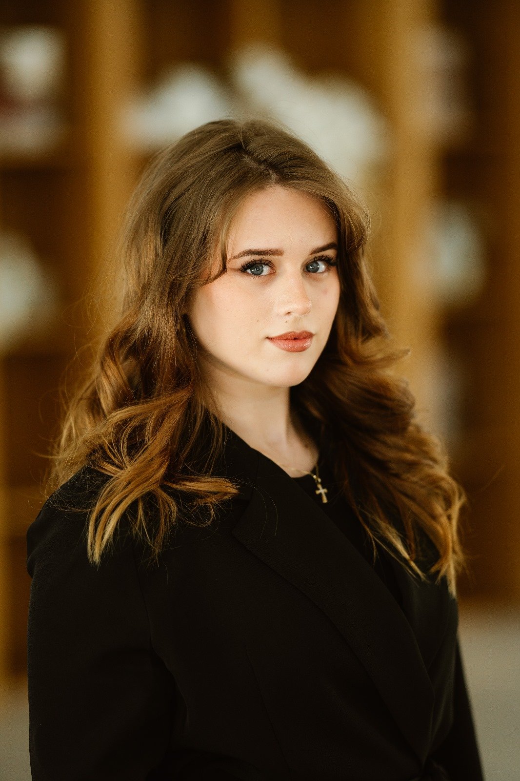 Portret Cătălina Anghel, în sacou negru, cu părul șaten ondulat, pe fundal neclar cu rafturi de bibliotecă