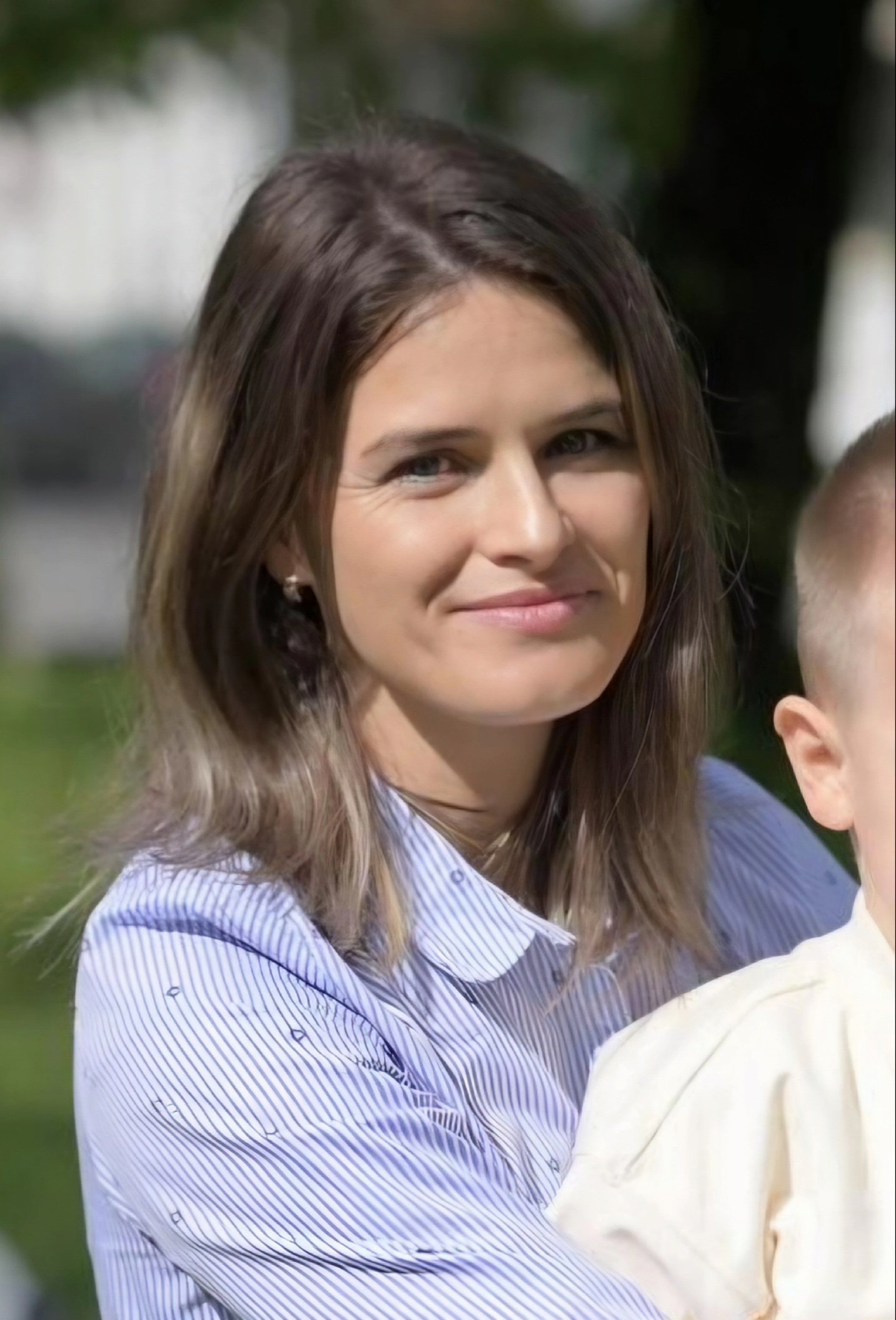 Portret Gabriela Cucea Cătană în cămașă cu dungi, ținând în brațe un copil întors cu spatele